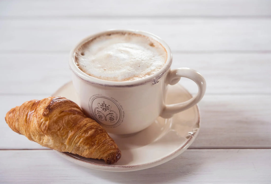 A cup of cappuccino with a croissant on a white table