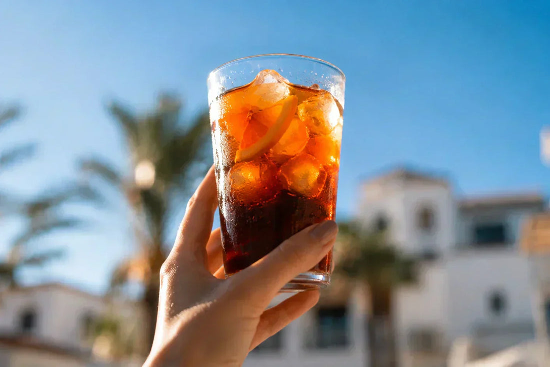Szklanka cold brew z lodem trzymana w dłoni na tle nadmorskiej scenerii z palmami i białymi budynkami – letni klimat JAVA Coffee.