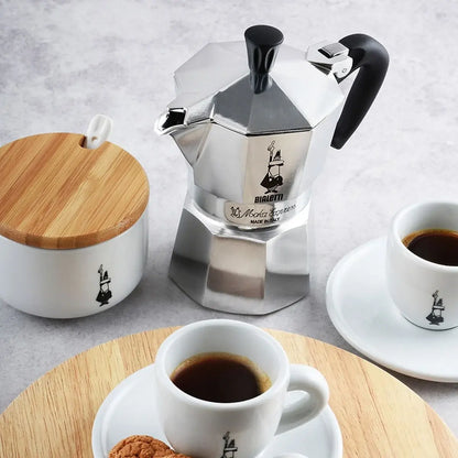  Kawiarka Bialetti Moka Express z filiżankami espresso i cukiernicą – włoski zestaw do parzenia kawy na tle jasnego blatu.