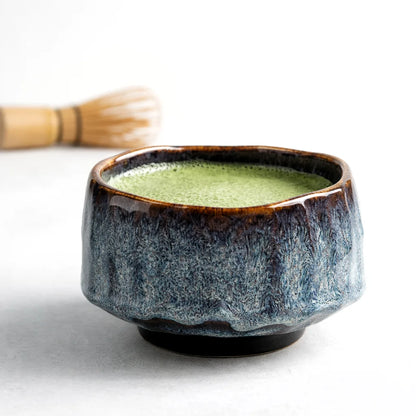 Ręcznie wykonana ceramiczna czarka do matchy z niebiesko-brązową glazurą i spienioną matchą na jasnym, minimalistycznym tle.