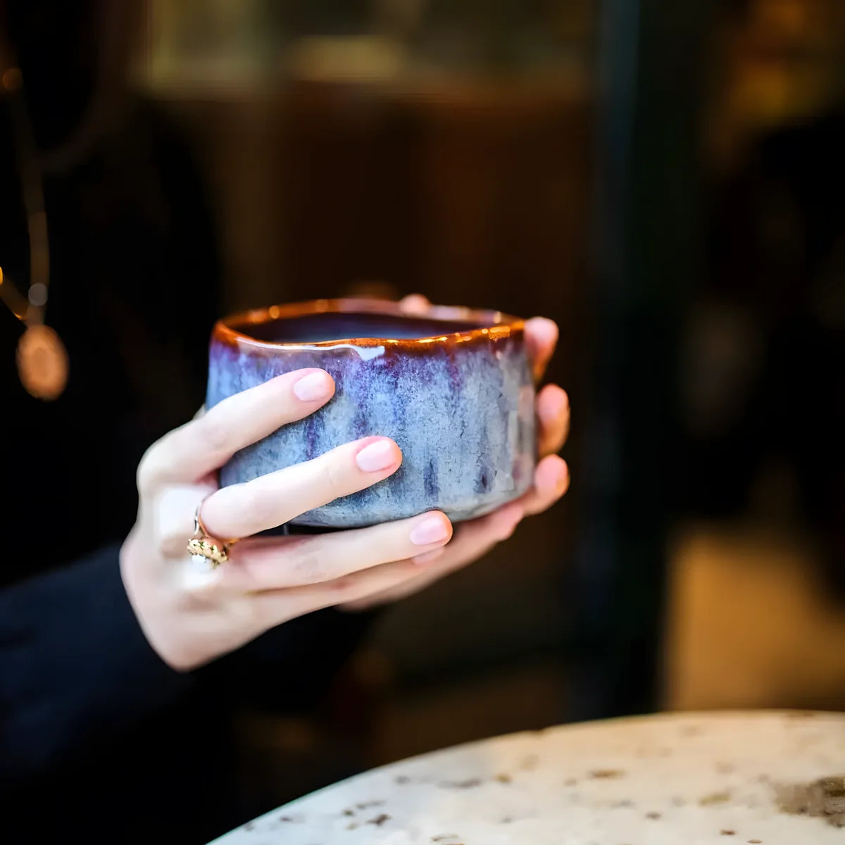 Ceramiczna czarka do matchy w niebiesko-brązowej glazurze trzymana w dłoniach, ręcznie wykonana miseczka do herbaty matcha w klimatycznym wnętrzu.