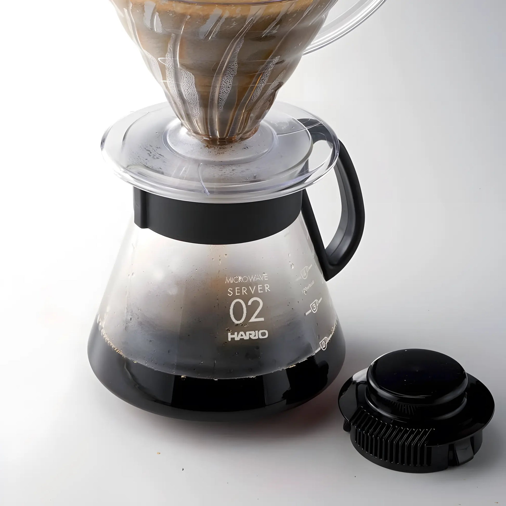 Serwer do kawy Hario Range Server V60 02 podczas parzenia, szklany dzbanek z zaparzoną kawą i dripperem V60, widoczna skala i pokrywka obok, zdjęcie produktowe
