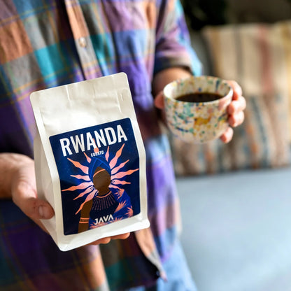 Osoba trzyma opakowanie kawy specialty Rwanda Cocatu od JAVA Coffee Roasters oraz filiżankę świeżo zaparzonej kawy. Zdjęcie przedstawia codzienny rytuał picia kawy specialty, eksponując opakowanie produktu i gotowy napar w domowym otoczeniu.
