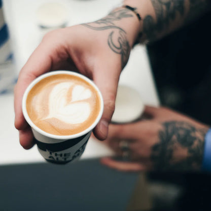  Kawa z mlekiem z latte art w kształcie serca w kubku jednorazowym – serwowana przez baristę z tatuażami na dłoniach, kawa specialty z mlekiem roślinnym.