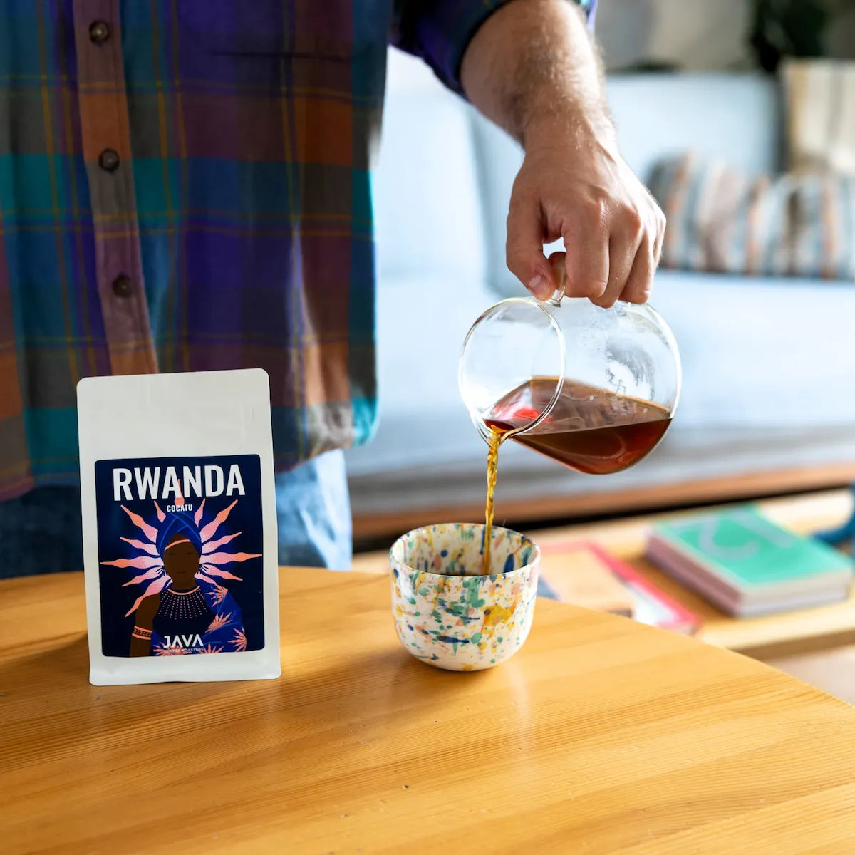 Przelewowa kawa specialty Rwanda Cocatu od JAVA Coffee Roasters nalewana z serwera do ręcznie wykonanej ceramicznej filiżanki. Na pierwszym planie opakowanie kawy Rwanda Cocatu, przedstawiające sposób serwowania i degustacji kawy specialty.
