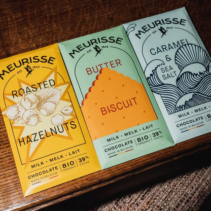 Trzy tabliczki mlecznej czekolady Meurisse: Roasted Hazelnuts, Butter Biscuit i Caramel & Sea Salt, ułożone na drewnianym stole w kolorowych opakowaniach z oznaczeniem BIO i Fairtrade.