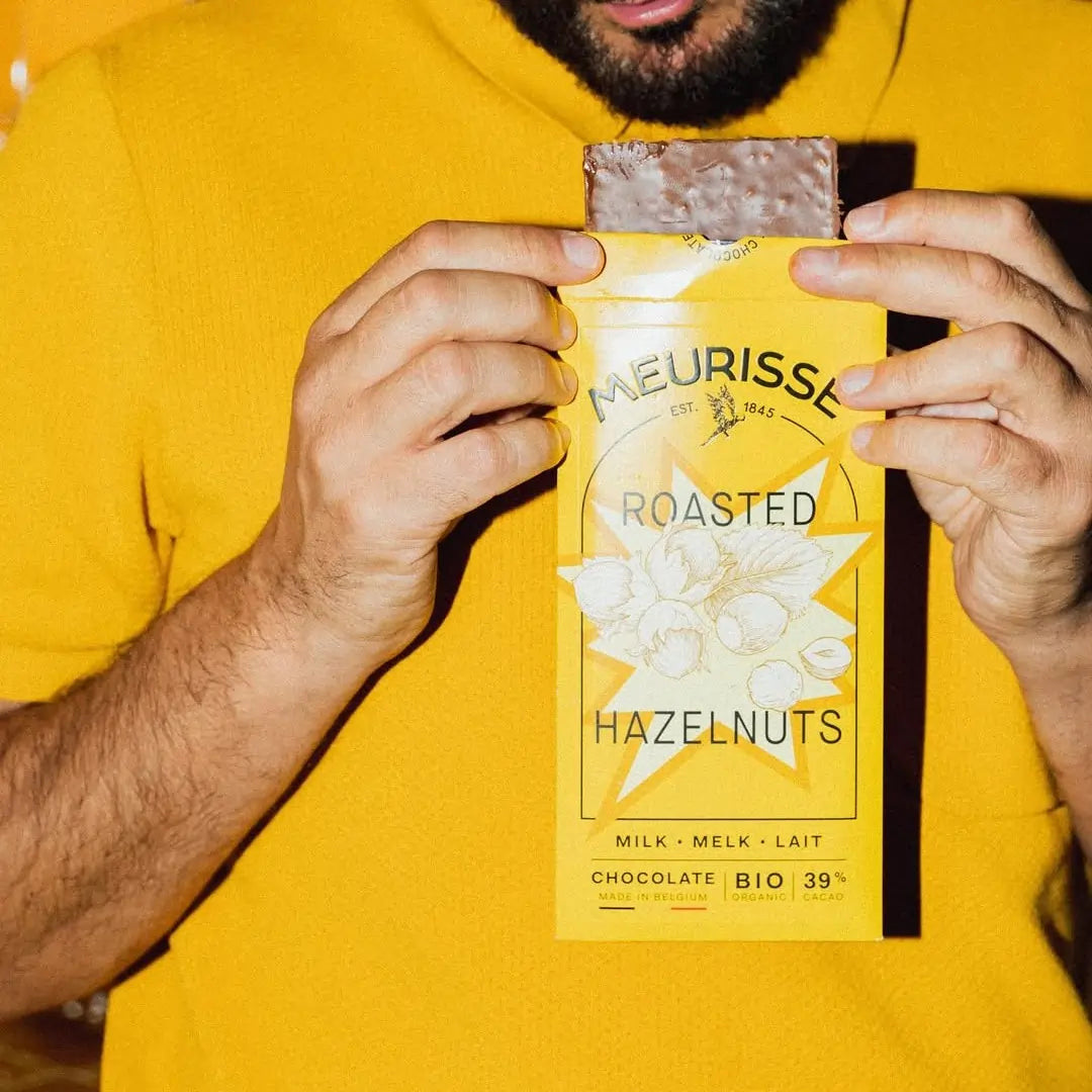 Osoba trzyma tabliczkę mlecznej czekolady Meurisse Roasted Hazelnuts 39% kakao w żółtym opakowaniu, dopasowanym kolorystycznie do ubrania, gotowa do degustacji.