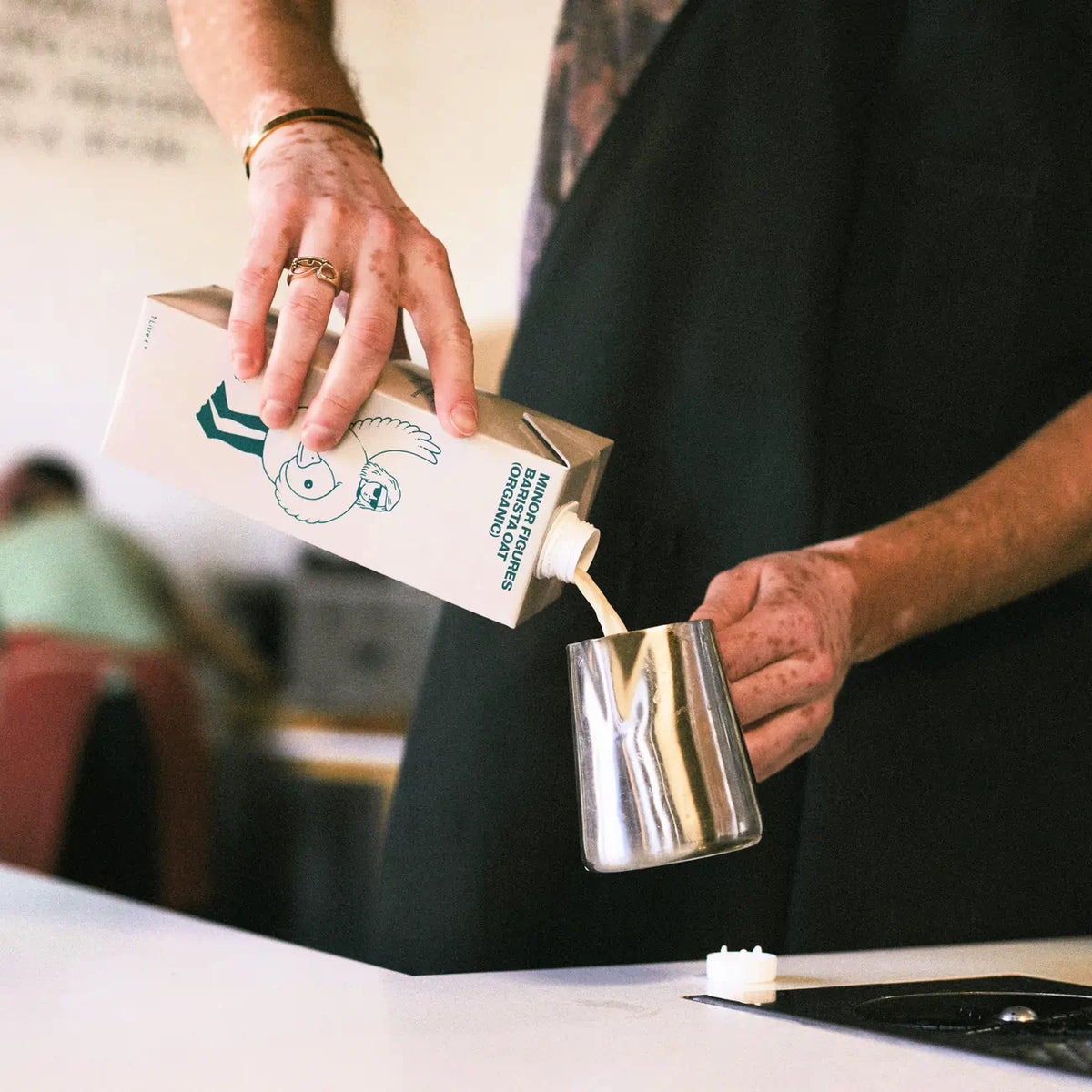 Barista nalewa napój roślinny Minor Figures Barista Oat Organic do dzbanka do spieniania – ujęcie z kawiarni, z widoczną osobą w tle i odkręconą zakrętką na blacie.