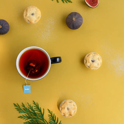 filiżanka herbaty Suki Tea Mince Pie z torebką herbaty i świątecznymi babeczkami mince pies na żółtym tle z dekoracjami z igliwia i figami