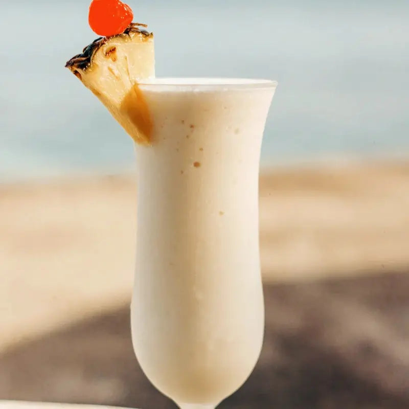  Egzotyczny drink litchi colada podany w wysokiej szklance, udekorowany kawałkiem ananasa i wisienką koktajlową, na tle plaży.