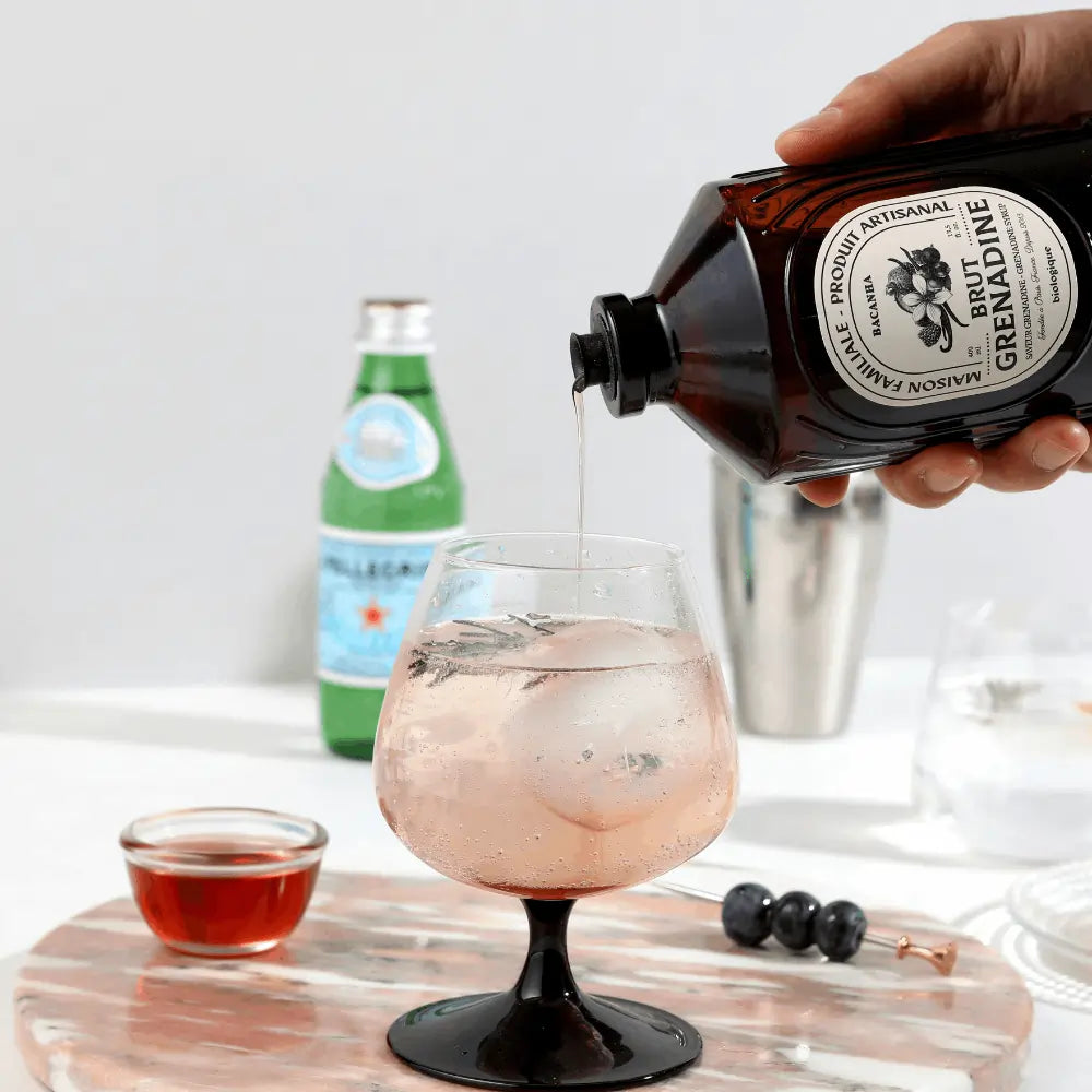Syrop Bacanha Grenadine w trakcie nalewania do szklanki z lodem – stylowe ujęcie przygotowania orzeźwiającego drinka z dodatkiem syropu o smaku czerwonych owoców i wanilii.