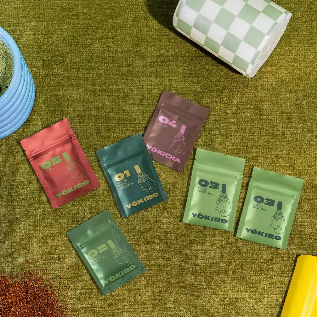 Lifestyle zdjęcie zestawu próbek Yōkiro – matcha ceremonialna, matcha traditional, hojicha i rooibos w kolorowych saszetkach na zielonym tle.
