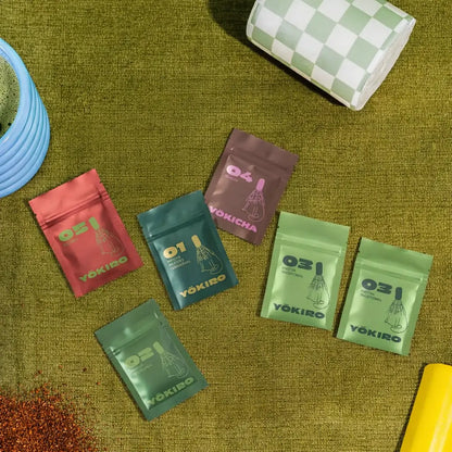 Lifestyle zdjęcie zestawu próbek Yōkiro – matcha ceremonialna, matcha traditional, hojicha i rooibos w kolorowych saszetkach na zielonym tle.